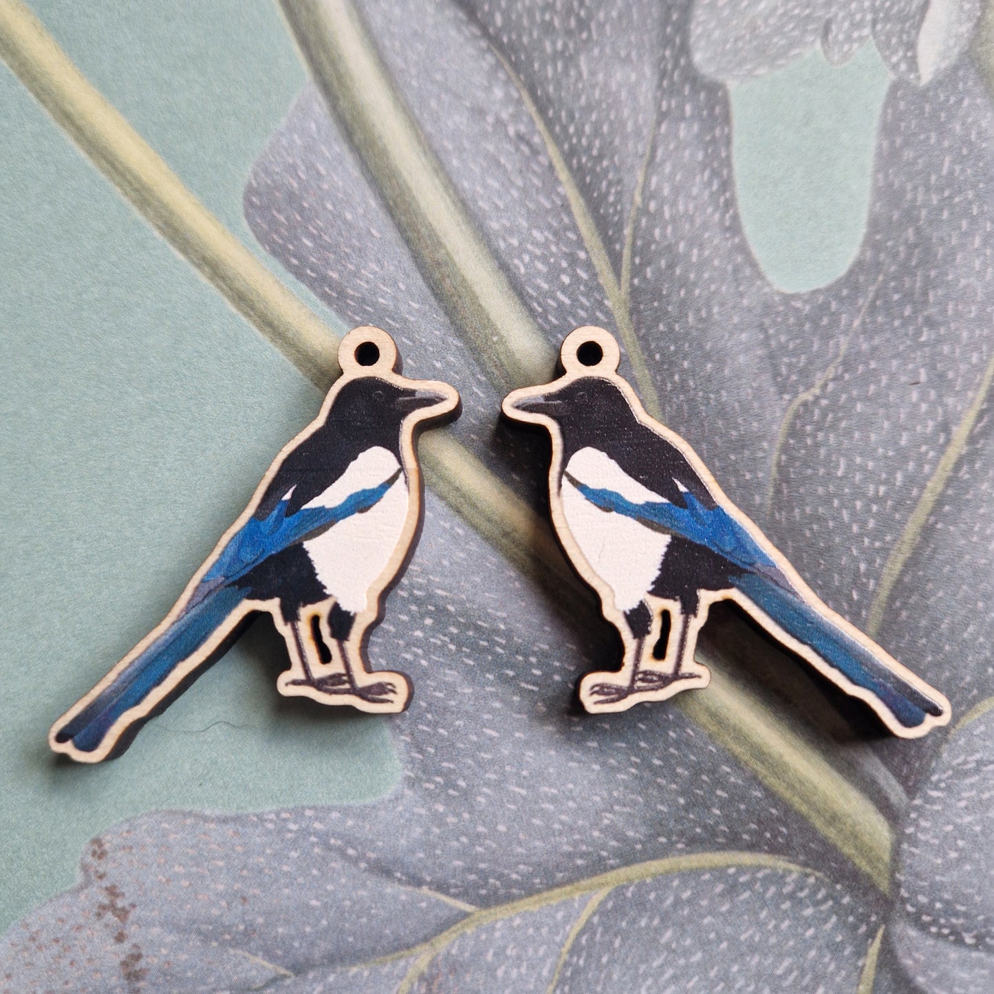 Two magpie-shaped wooden charms by Duck Egg Designs, featuring black, white, and blue markings, rest on a leafy pastel background. Each charm has a small top hole for hanging or creating unique Magpie Earrings.