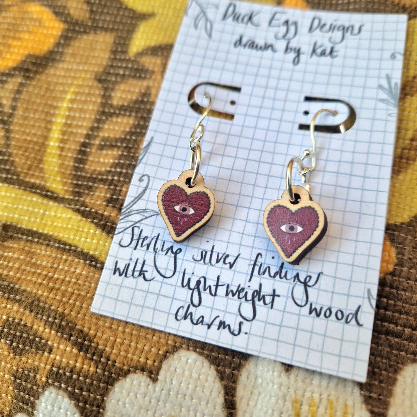 Mini Heart Dangle Earrings by Duck Egg Designs, featuring an eye motif on lightweight wood with sterling silver findings, displayed on a card with handwritten text against a patterned fabric background.