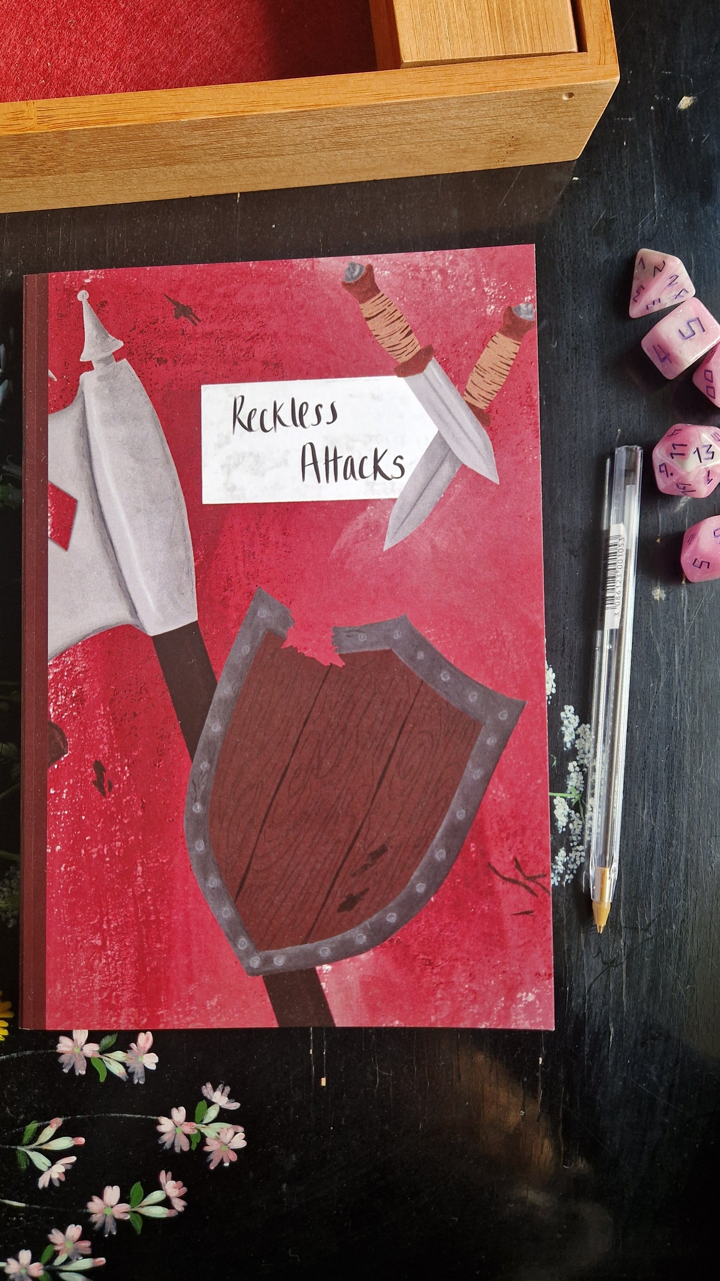 The Ways Of The Wood Notebook by Duck Egg Designs is a red A5 lined notebook with illustrations of a shield, battle axe, and daggers. Purple dice, a pen, and a wooden box sit next to this recycled paper notebook on a dark table.