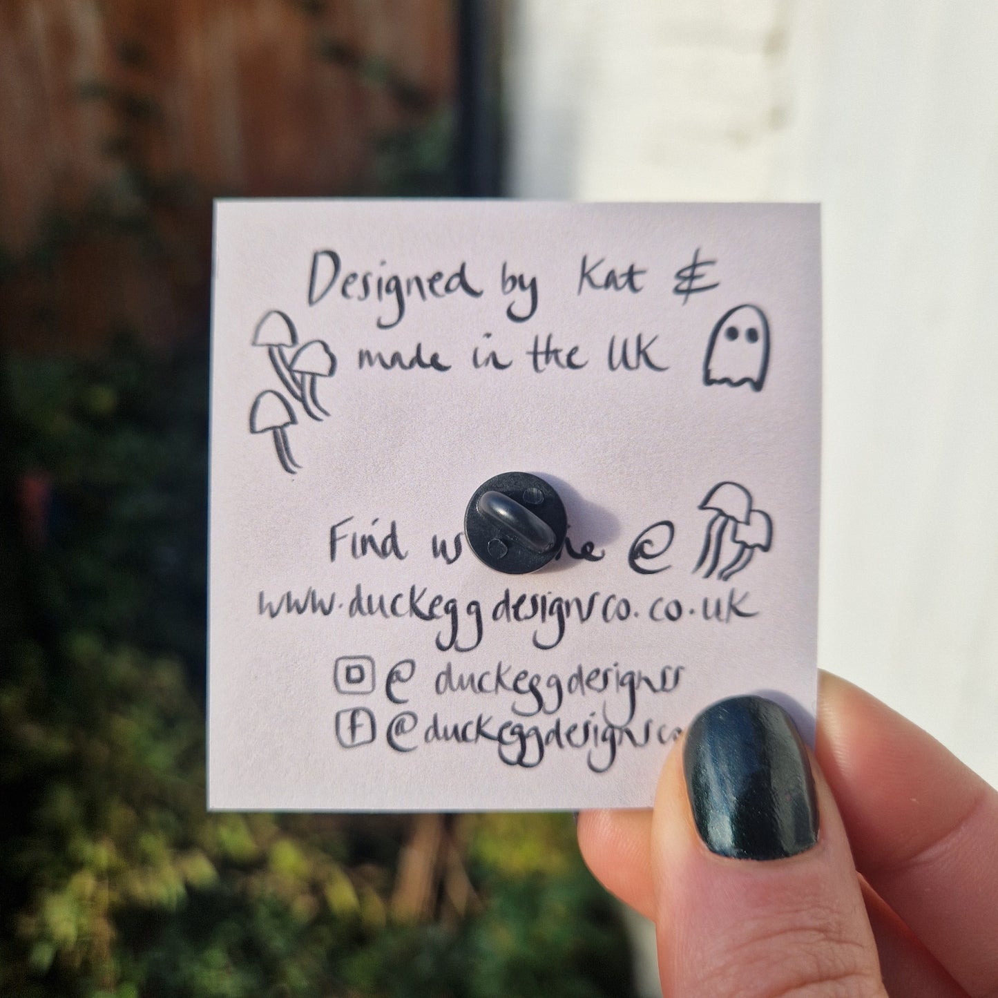 The back of the pin badge, showing the black rubber pin back on the dusky pink of the backing card. The backing card is held by a white hand wearing black nail polish and featuring the words 'Designed by Kat & made in the UK' with website and social media information below.