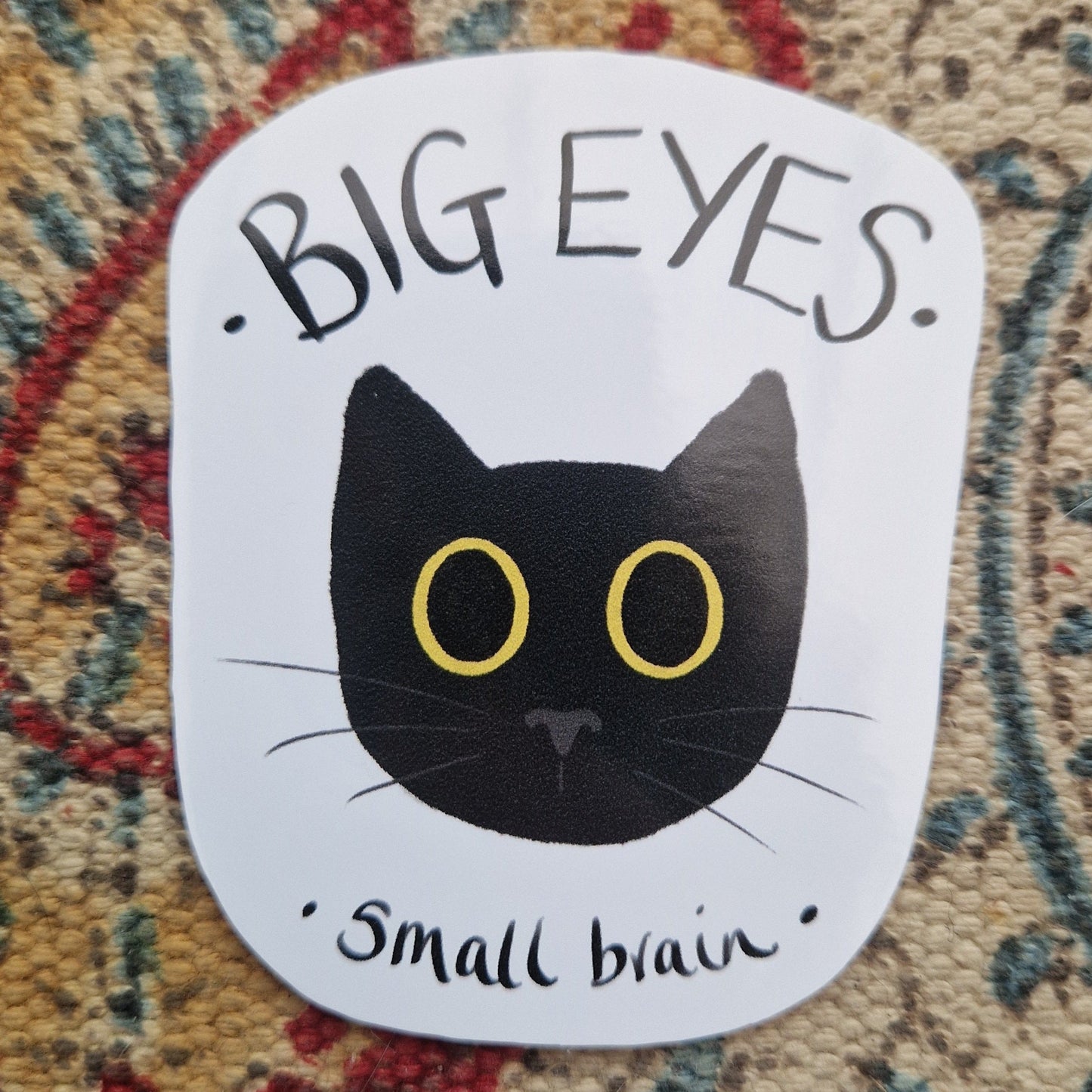 A closer look at the sticker showing the detail in the cat head and the handwritten text. Behind the sticker is a patterned fabric background.