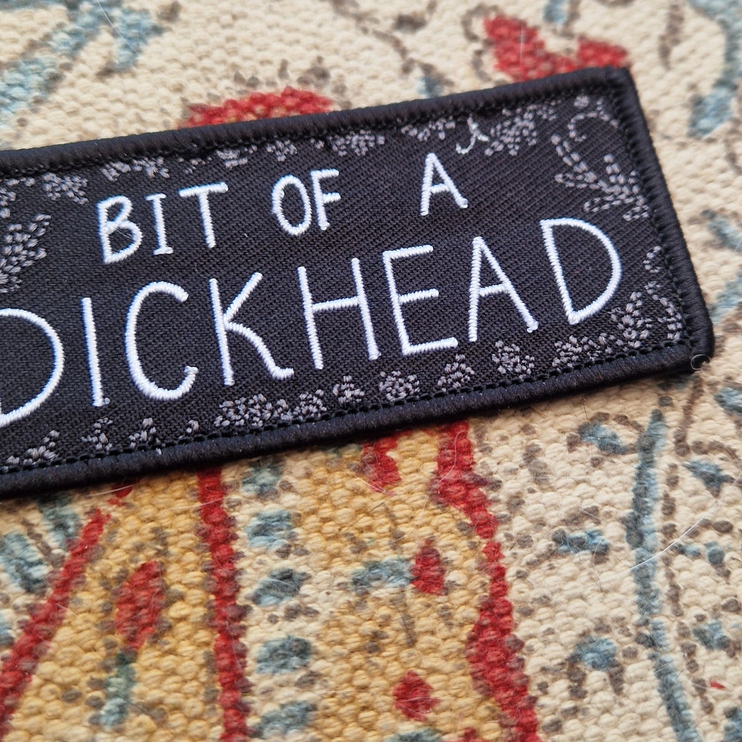 A black rectangular patch with the words 'BIT OF A DICKHEAD' across the middle in white text and a grey leafy frond design around the outside. The patch sits on a patterned fabric background.