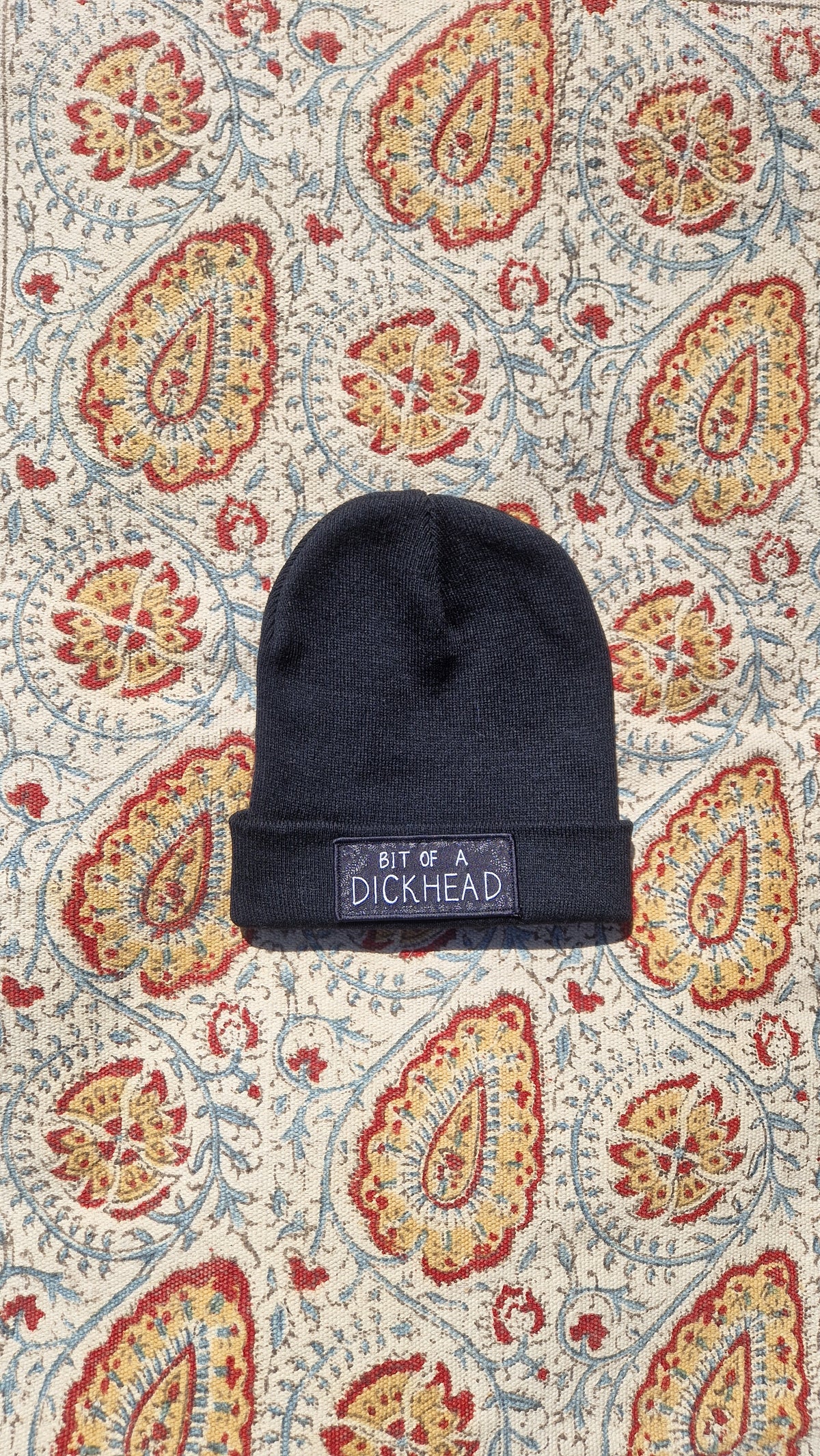 A black beanie lays on a patterned fabric background. The beanie has a rectangular patch on it that reds 'Bit Of A Dickhead'.