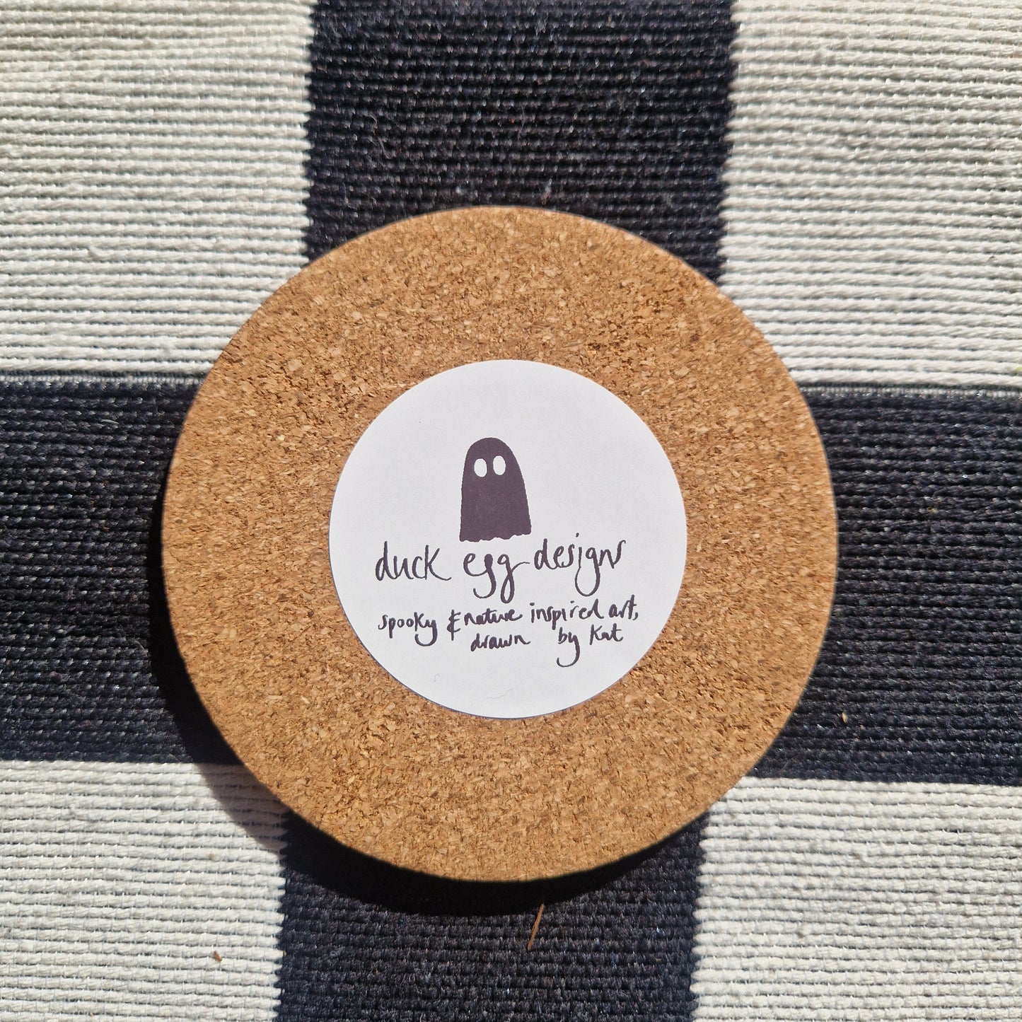 A round cork coaster base with a white circular sticker with the ghost duck egg designs logo on it. The coaster is on a checkered white and black fabric.