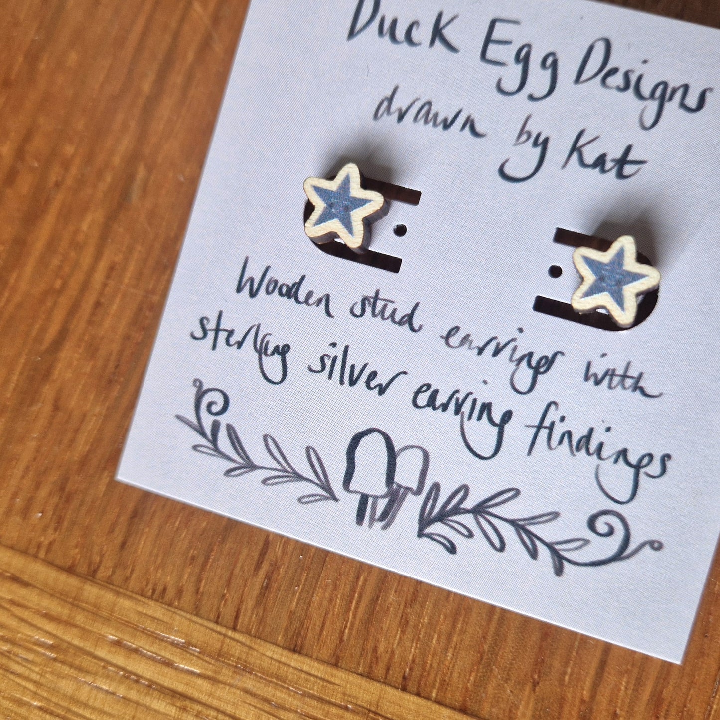A close up of the pair of grey star earrings with small smiling faces sit on a light purple backing card with the words 'Duck Egg Designs drawn by Kat' across the top in black and 'Wooden stud earrings with sterling silver earring findings' across the bottom. Behind the backing card you can see a wooden background.