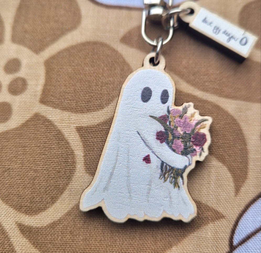 The Ghost Flower Keyring by Duck Egg Designs is a UK-made, sustainable wood keychain shaped like a cute spirit holding flowers, with a red heart on its chest. It features brown and beige floral-patterned fabric.