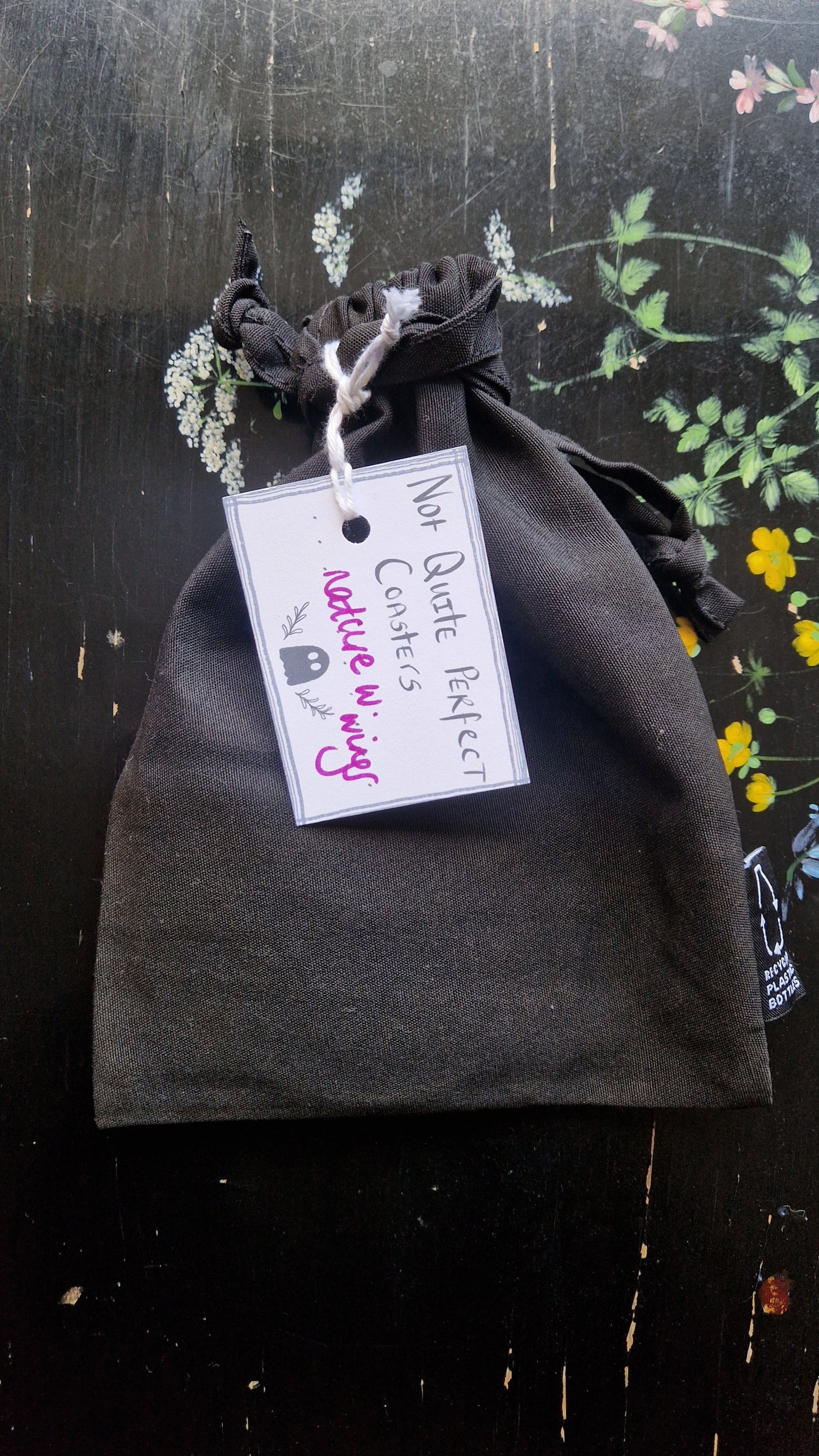 A black fabric drawstring pouch sits on a black table with flowers painted on it. The pouch has a white label that reads 'Not Quite Perfect Coasters' in black writing with the words 'nature w. wings' in purple written on it.
