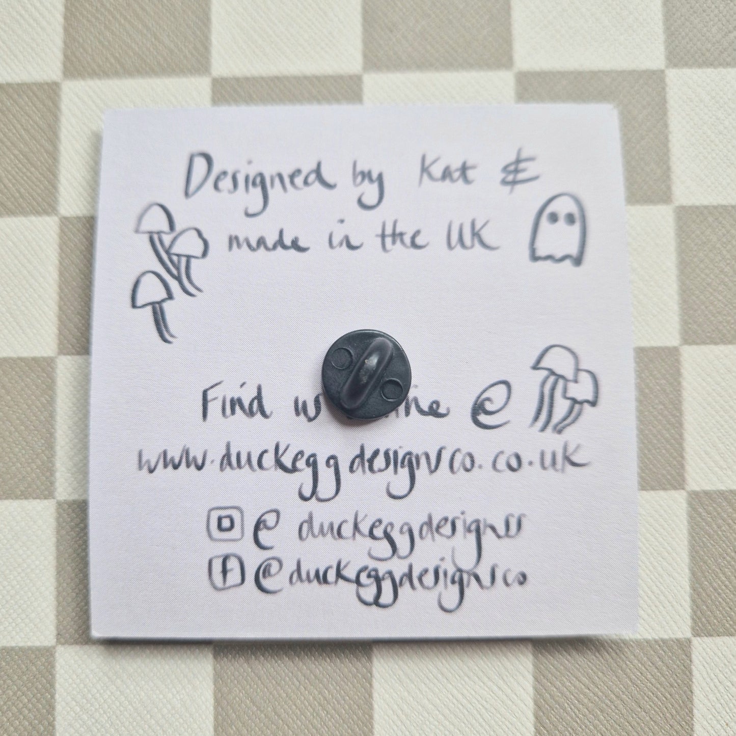 The back of the pin badge showing the rubber back as well as the reverse of the pin badge backing card which is a dusky lilac colour and black handwritten information about the pin.