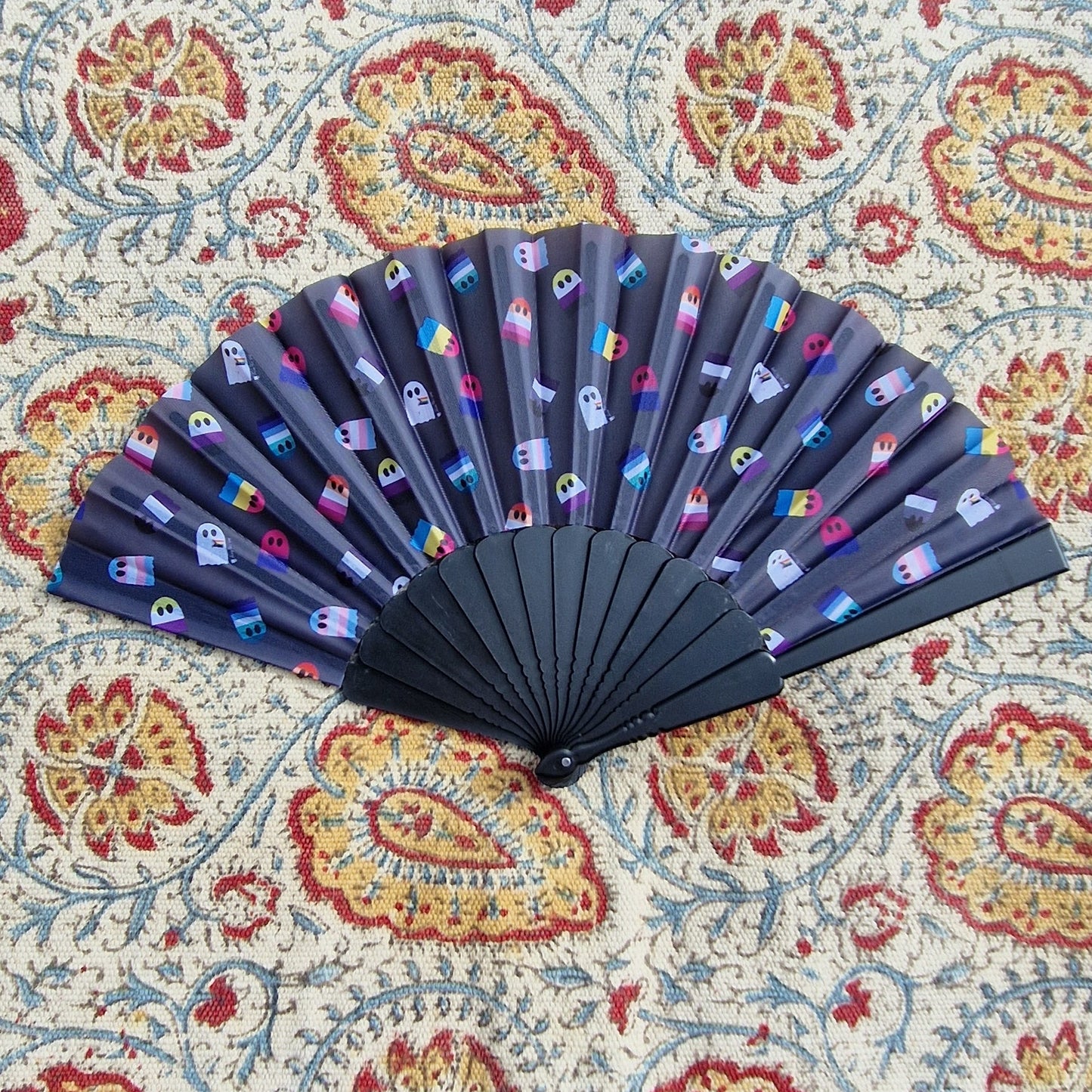 A black fan sits on a patterned rug. The fan has a repeat print of different pride flags as ghosts.