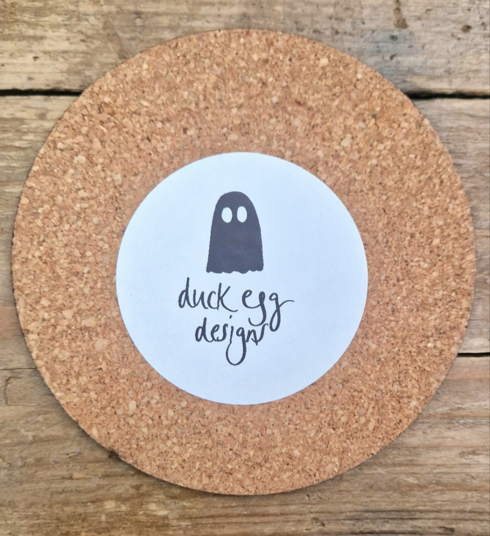 The base of the round coaster showing the cork bottom featuring a white round sticker with the black ghost duck egg designs logo.