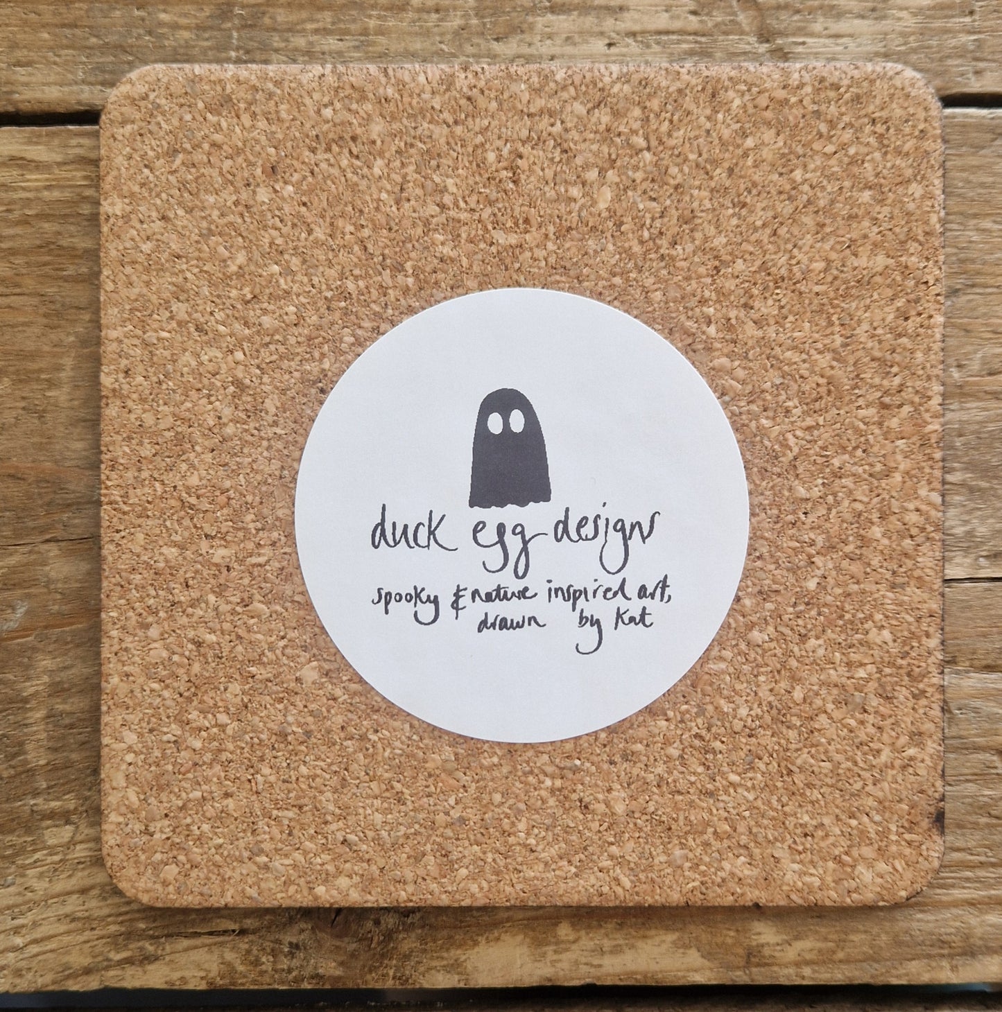 The back of the cork coaster showing a round white sticker with the ghost Duck Egg Designs logo. Behind the coaster is a wooden planks background.