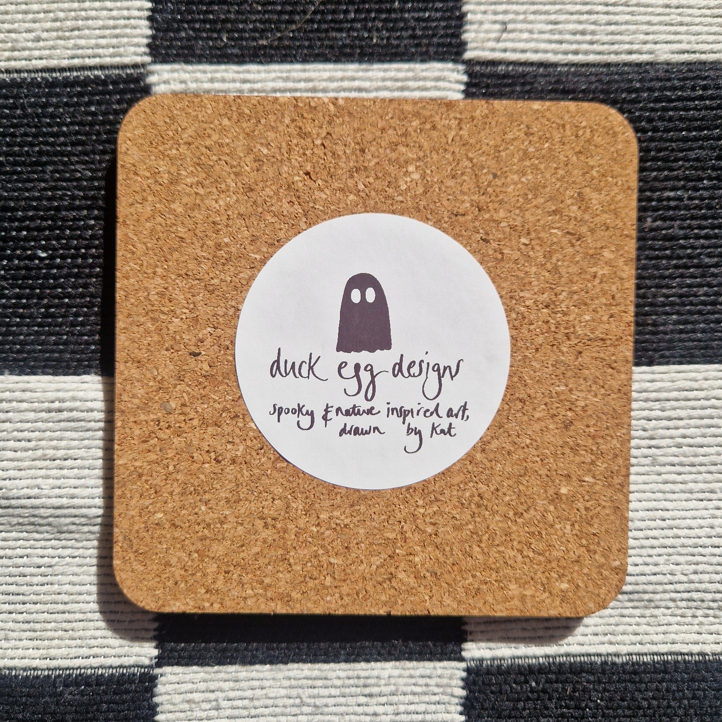 The back of a square coaster showing the cork base with a white round sticker featuring the ghost Duck Egg Designs logo.