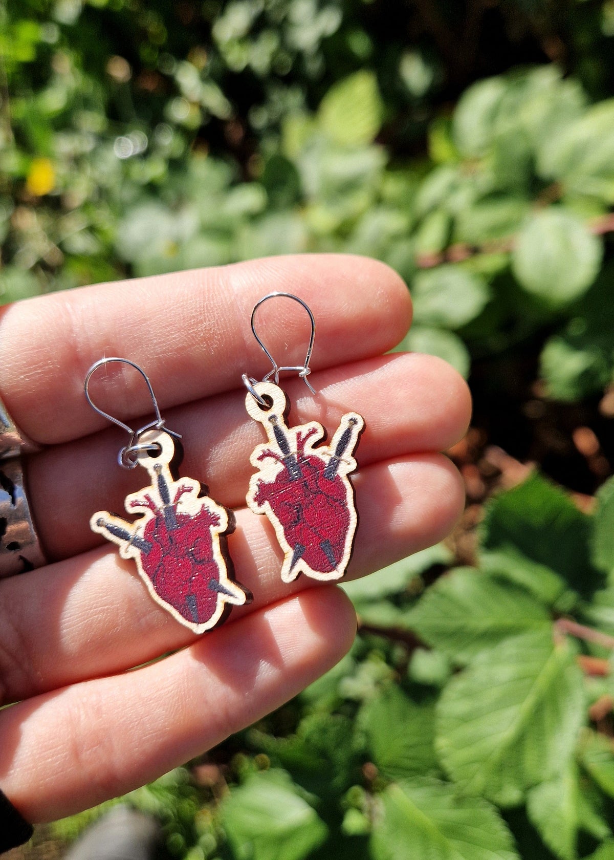 A pair of red, black and grey anatomical heart and dagger earrings with sterling silver earring findings sit on a white hand wearing silver rings above an out of focus patch of brambles.