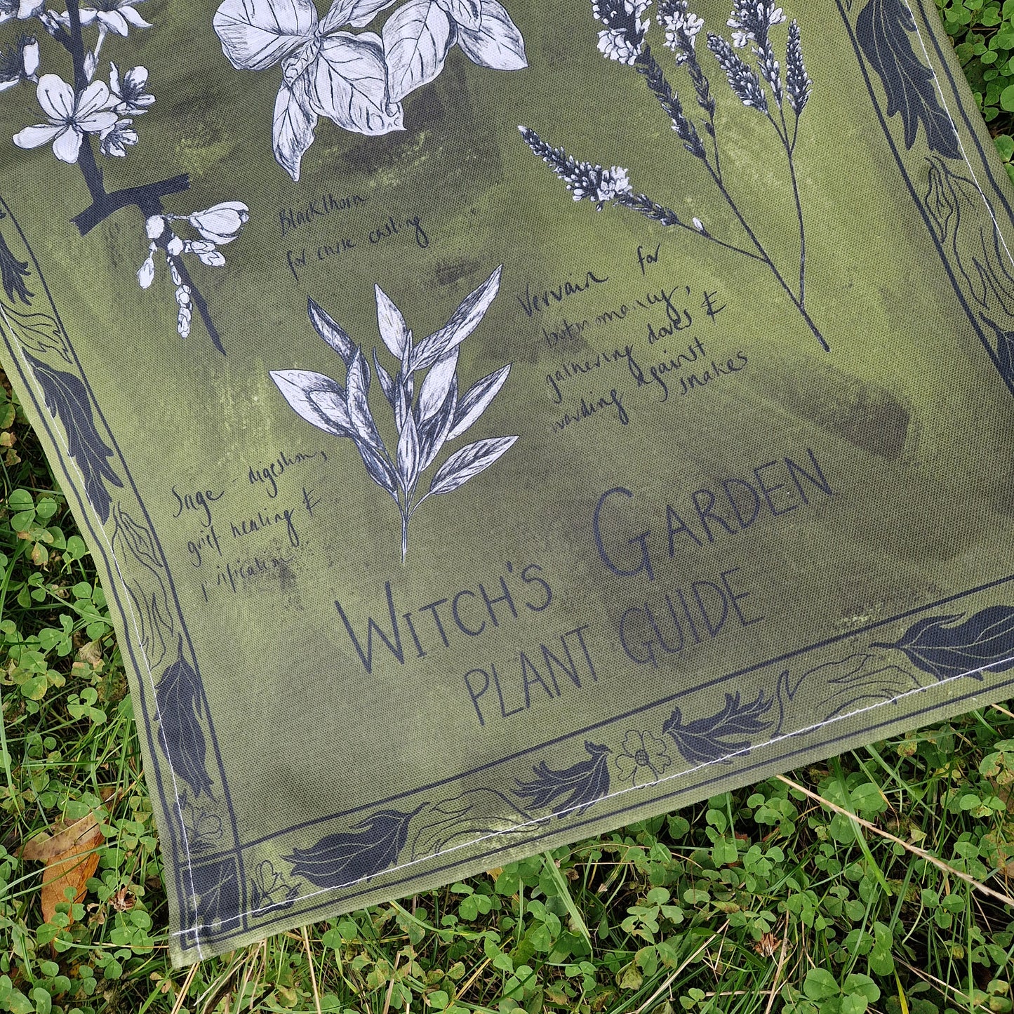 The bottom section of the mottled green tea towel, showing the words 'witch's Garden Plant Guide' in black handwriting as well as the plant filled border design surrounding the detailed botanical planbt illustrations. The tea towel sits on a patch of clover.