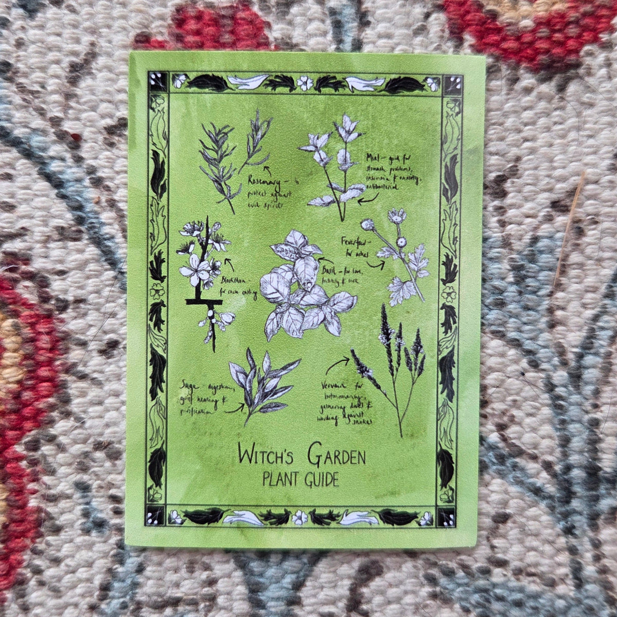 A rectangular green sticker with different plants on that you may find in a witchs garden illustrated in black and white with a black border. Behind the sticker is a patterned fabric background.