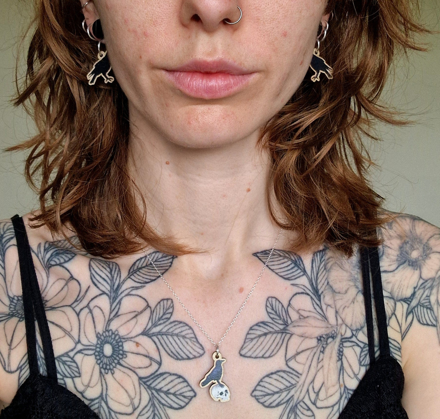 Woman with floral tattoos on her shoulders wearing black raven earrings and a crow on a human skull necklace.