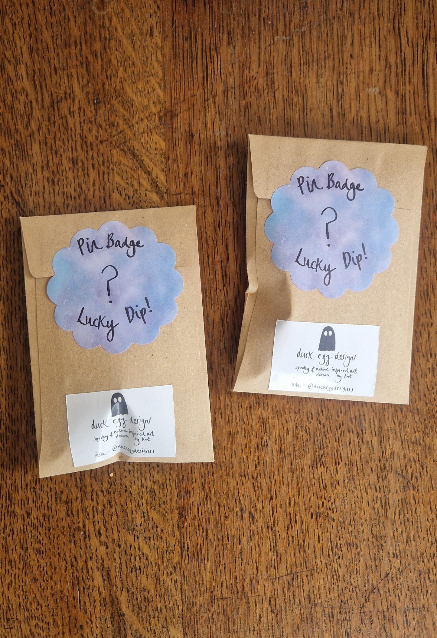 Two brown paper packets labeled "Mystery Pin Badge" by Duck Egg Designs with pastel cloud stickers and white business cards on a wooden surface. Each packet contains a surprise enamel pin from Duck Egg Designs.