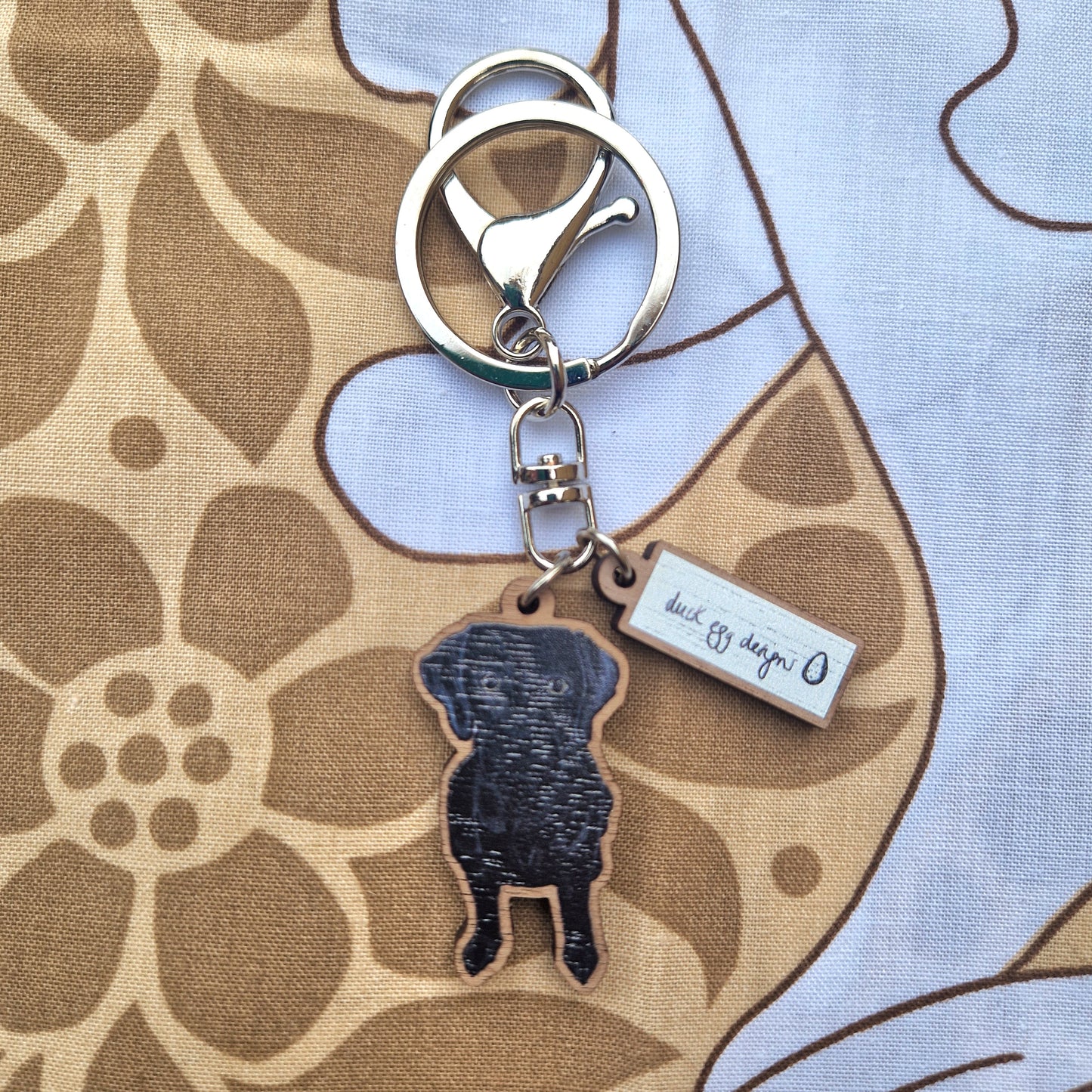 The Rondo Black Labrador Keyring by Duck Egg Designs is crafted from FSC certified wood, featuring a black labrador silhouette and a branded tag, displayed on brown and beige floral fabric.