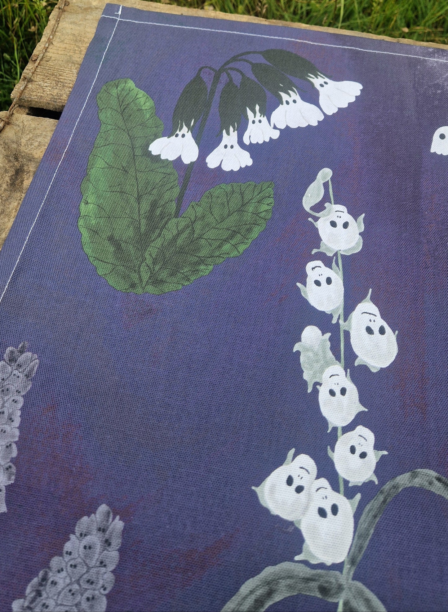 The Spooky Flowers Tea Towel by Duck Egg Designs features a whimsical botanical print with white flowers, green leaves, and ghost-like faces on a purple background—an enchanting addition to any kitchen.