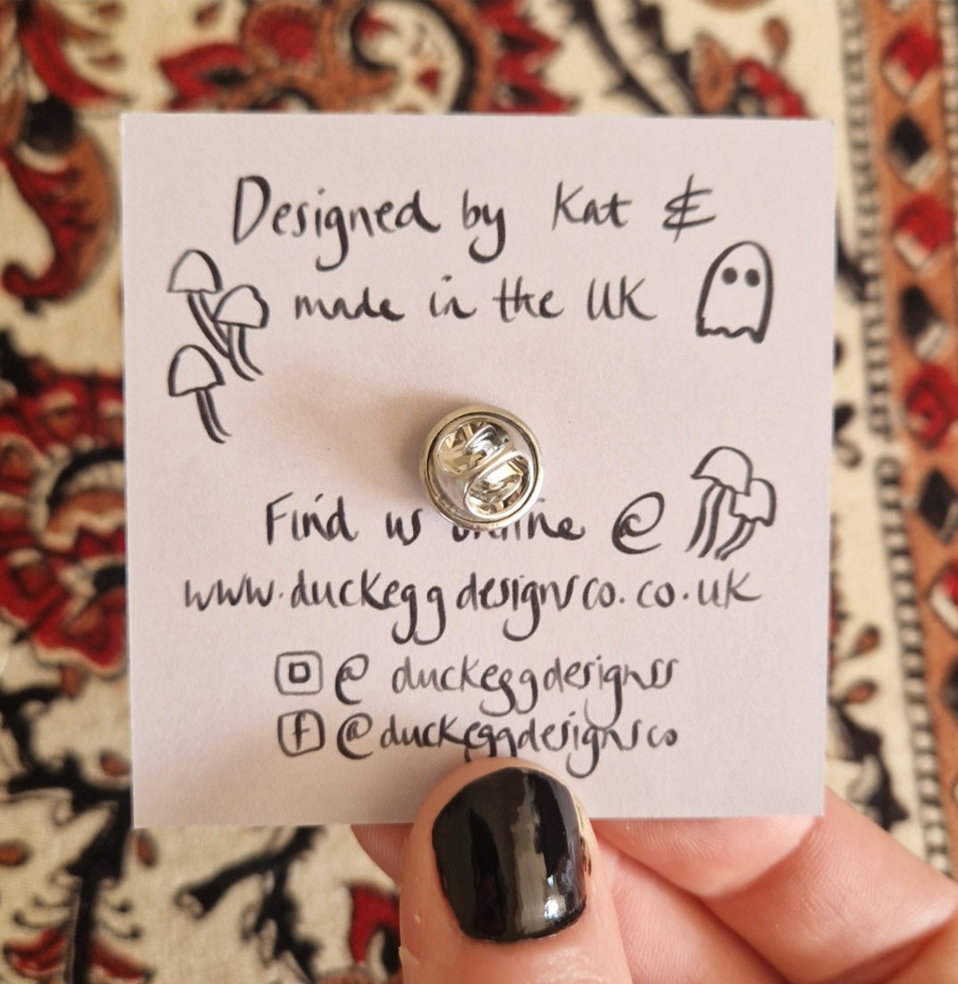 A hand with black-painted nails holds a card reading "Designed by Kat & made in the UK," adorned with mushroom and ghost doodles. The Stan Pigeon Mini Pin by Duck Egg Designs, crafted from FSC-certified wood, rests beside social media info on patterned fabric.