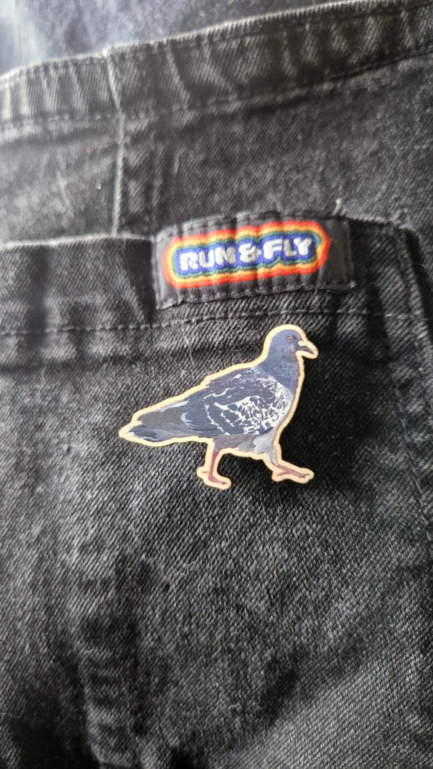 The Cyril Pigeon Pin Badge by Duck Egg Designs, made in the UK, is pinned to black denim jeans. Above it, a patch featuring RUN & FLY in colorful letters is sewn onto the pocket.