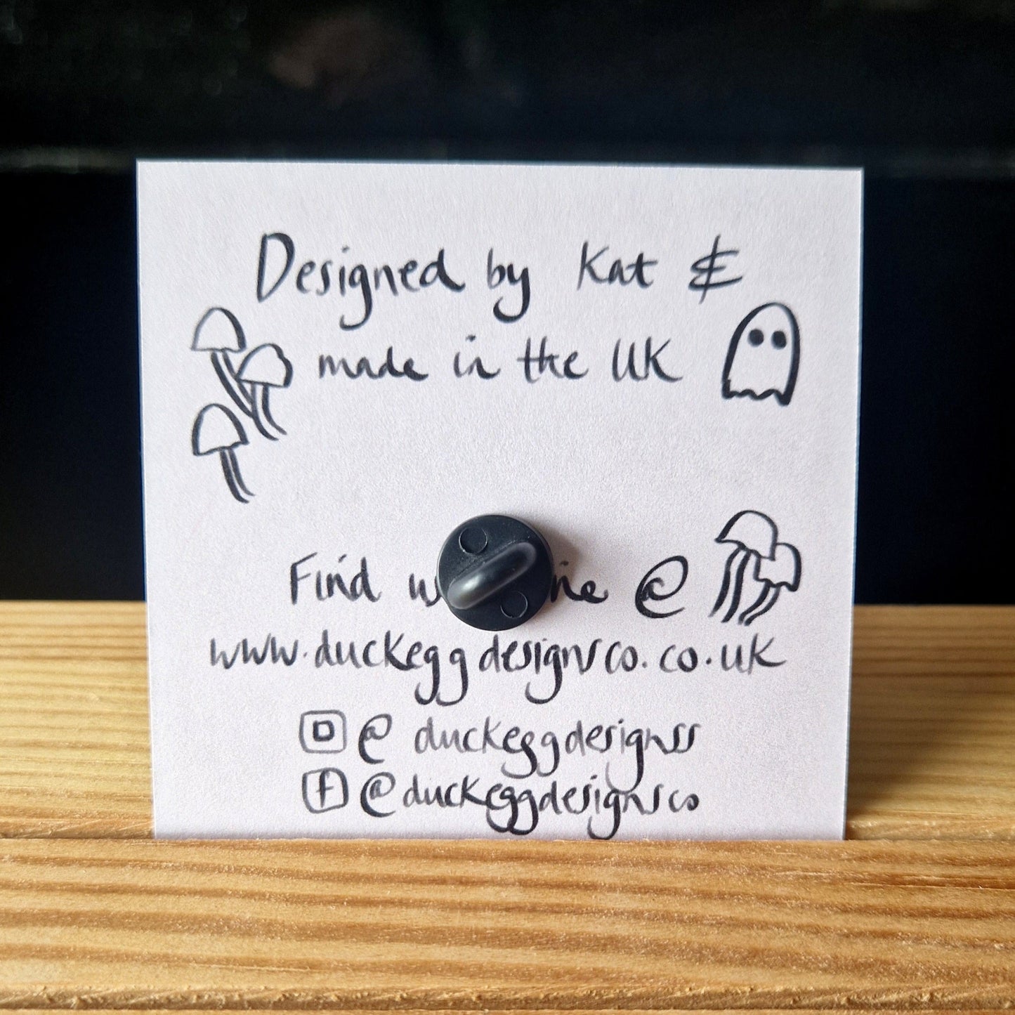 A handwritten note with mushroom and ghost doodles says: Designed by Kat & made in the UK. Find my Badger Skull and Wildflowers Pin Badge @ www.duckeggdesignco.co.uk, or follow Duck Egg Designs on Instagram and Facebook. A button is attached.