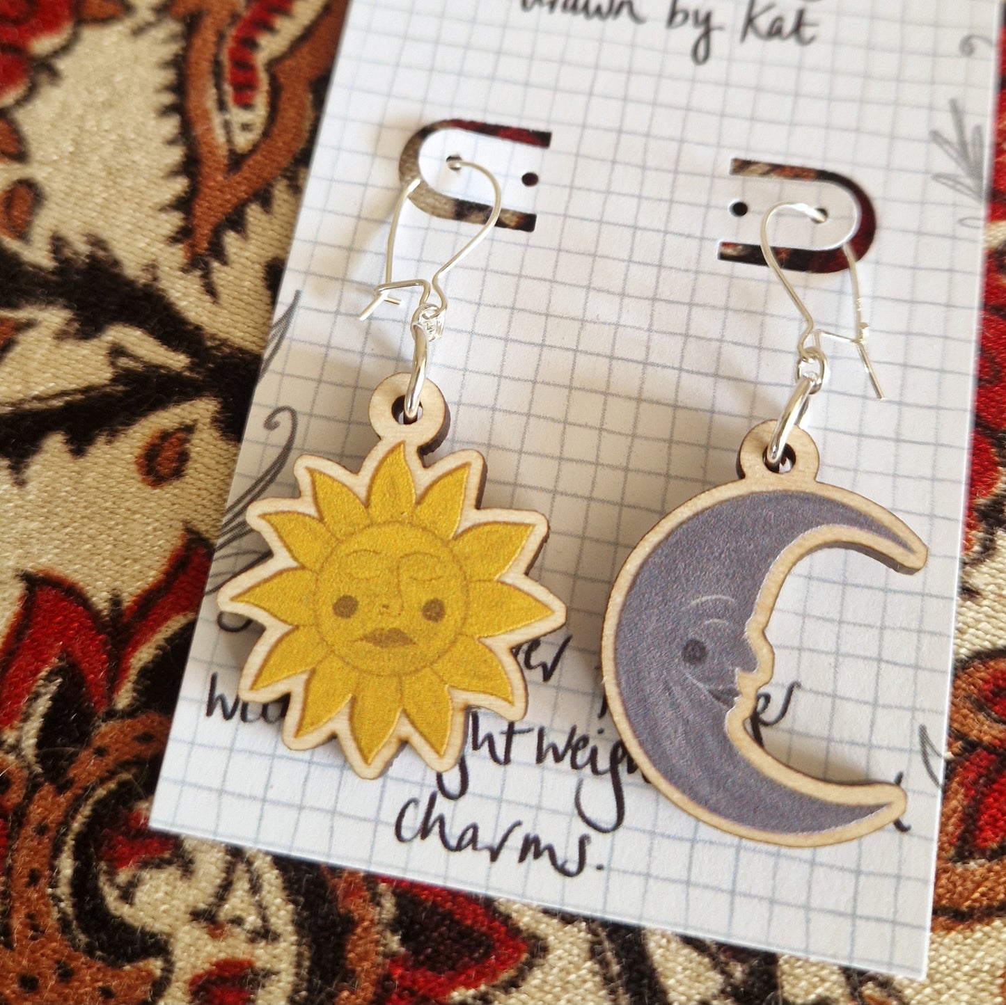 Duck Egg Designs Sun and Moon Face Earrings—wooden pieces featuring a smiling yellow sun and gray crescent moon—are displayed on a checkered card atop patterned fabric.