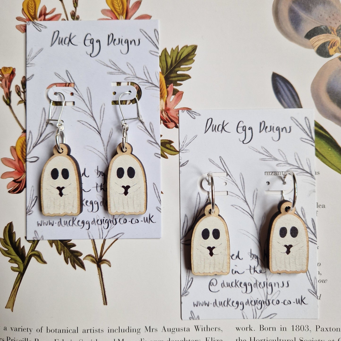 Two pairs of Duck Egg Designs' Ghost With Heart Earrings with cute faces are displayed on branded floral cards, set against a background of botanical illustrations and partial book text.