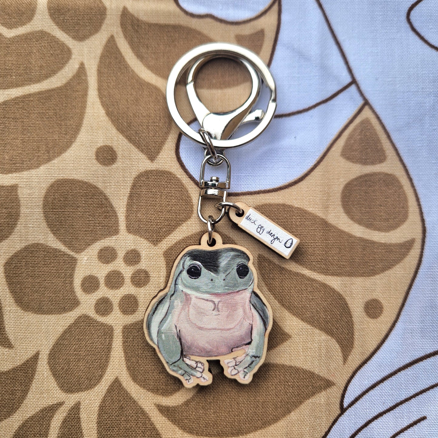 The Duck Egg Designs Frog Keyring features a cute cartoon frog charm and a small "but its you :)" tag, displayed on a brown floral-patterned fabric background.