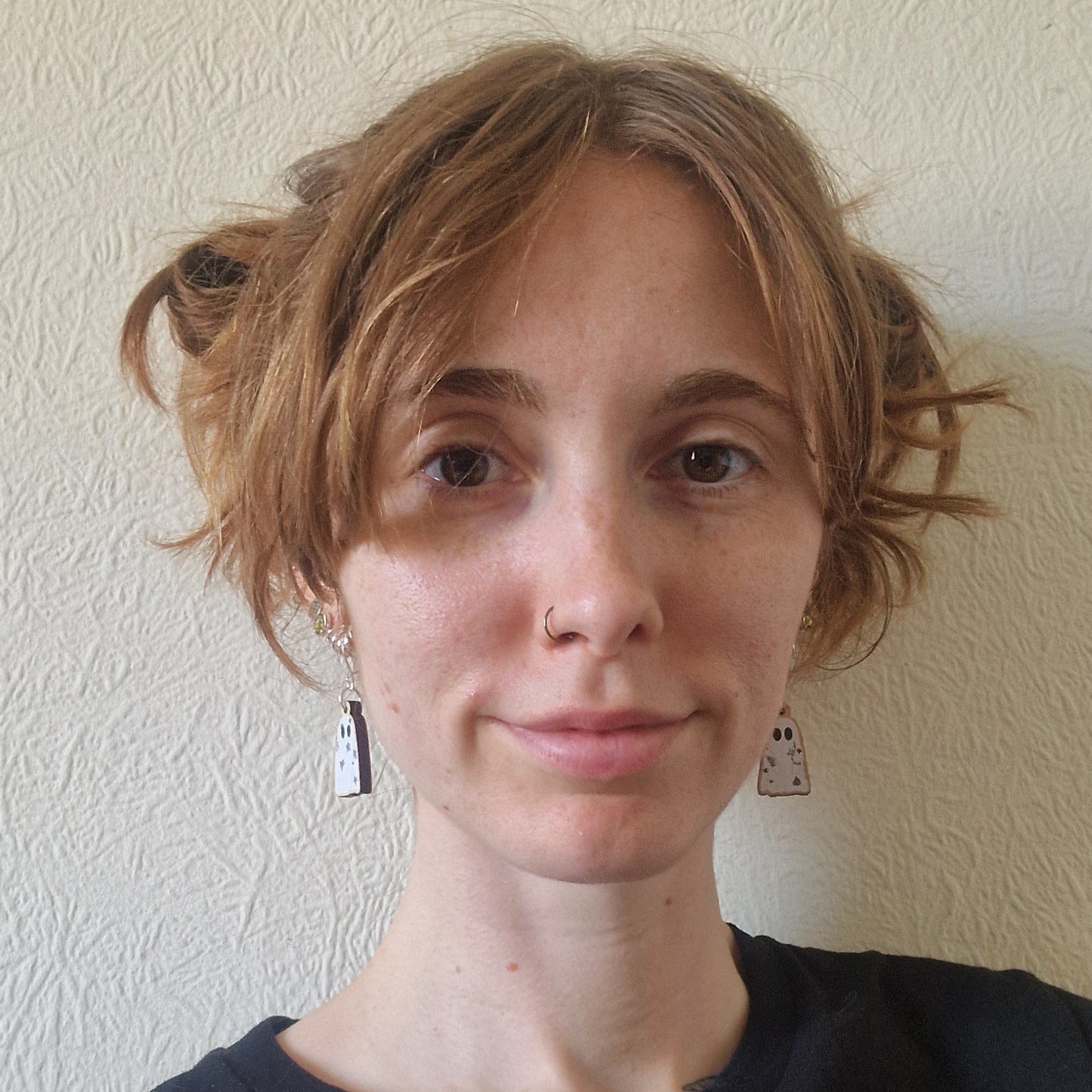 A young person with short, wavy light brown hair in pigtails, wearing a black shirt and Duck Egg Designs Tattooed Ghost Earrings, stands smiling softly at the camera against a textured beige wall.