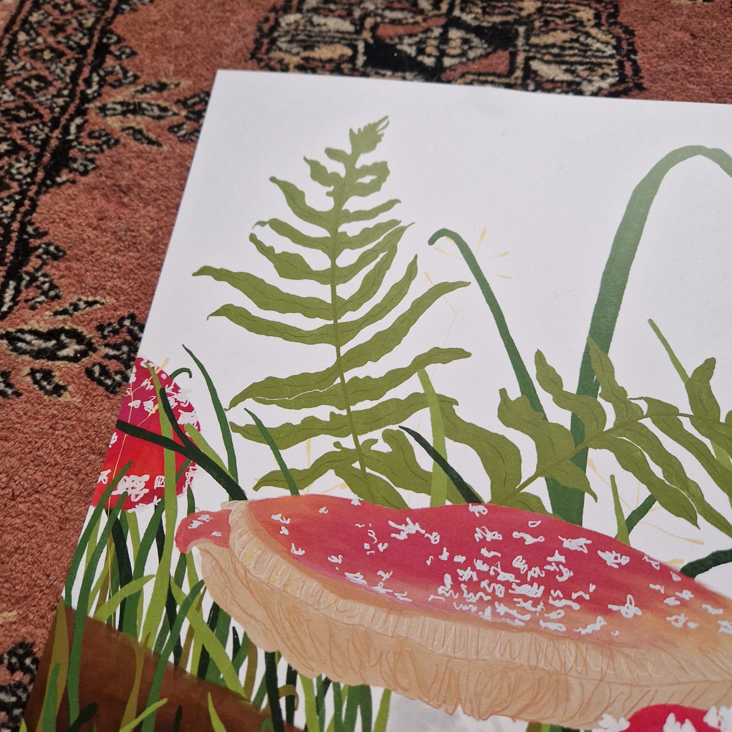 A close up of the top left of the print showing the top of the main fly agaric fungi as well as the green grasses and ferns on the white background. Above and to the left of the print you can see a red textured patterned background.