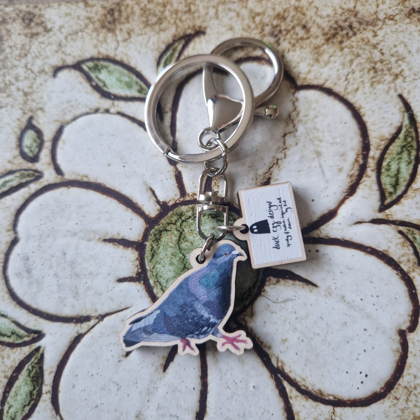 The Paloma Pigeon Keyring by Duck Egg Designs features a pigeon charm and a small rectangular tag, shown resting on ceramic with a white flower and green leaves.
