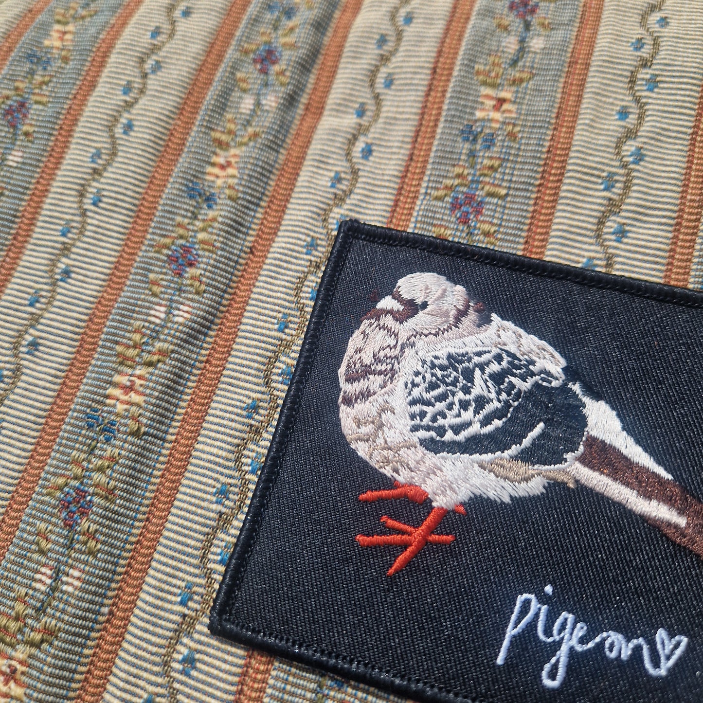 The Embroidered Bert Pigeon Patch by Duck Egg Designs is a square black patch with a pigeon and heart, plus the word pigeon, shown on striped floral fabric. Add a whimsical touch to your projects with this charming design.
