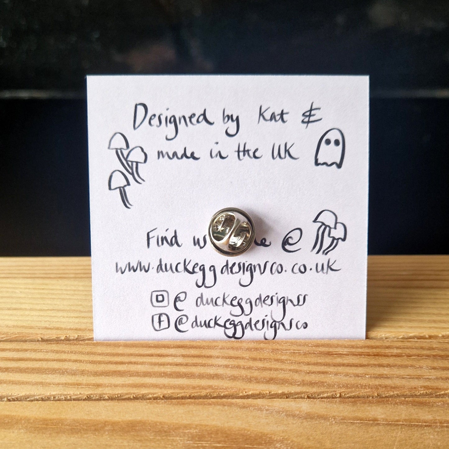 A square card with a silver pin fastener, featuring the Ghost Eating Chocolate Mini Pin. Illustrated by Kat for Duck Egg Designs and made in the UK, card includes social media and website details.