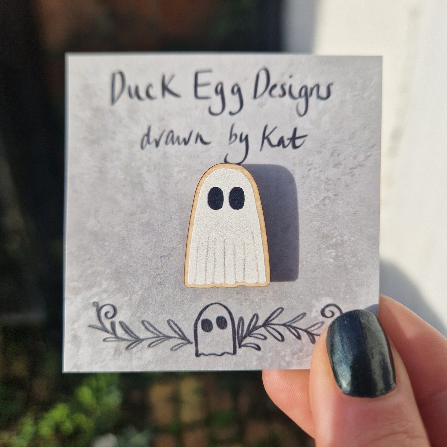 A white ghost pin badges made of wood attached to a grey backing card that reads "Duck Egg Designs drawn by Kat'. The backing card is being held in by a hand with black nail polish in front of a building.
