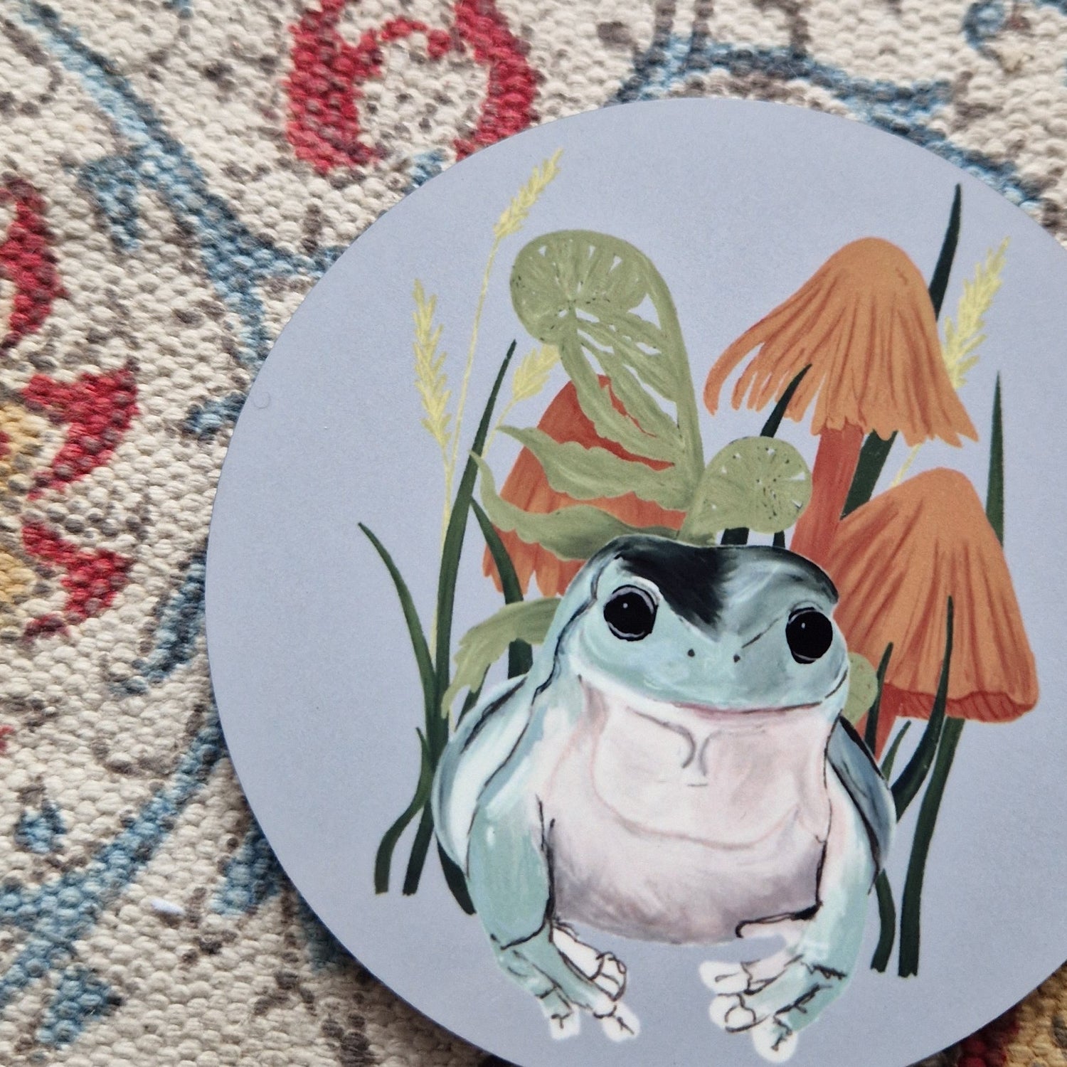 A round grey coaster with a friendly green frog, orange fungi and green ferns design. Behind the coaster is a white patterned fabric.