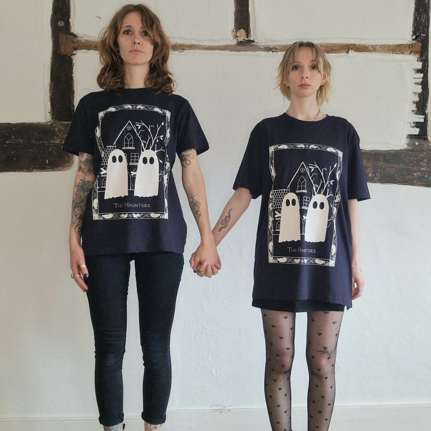 Two white women stand in front of a white wall with exposed wooden beams holding hands wearing dark blue tshirts with a ghost design. 