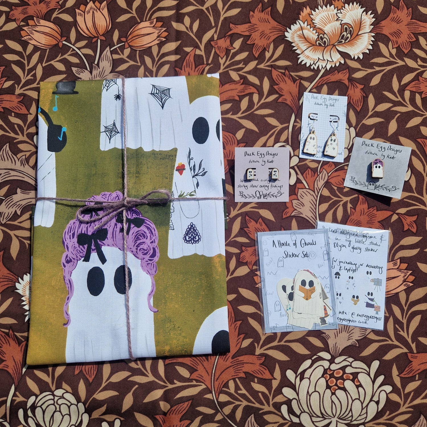A colourful orange ghost teatowel lies folded next to two ghost pairs of earrings, a pink wigged ghost pin badge and a ghost sticker set.
