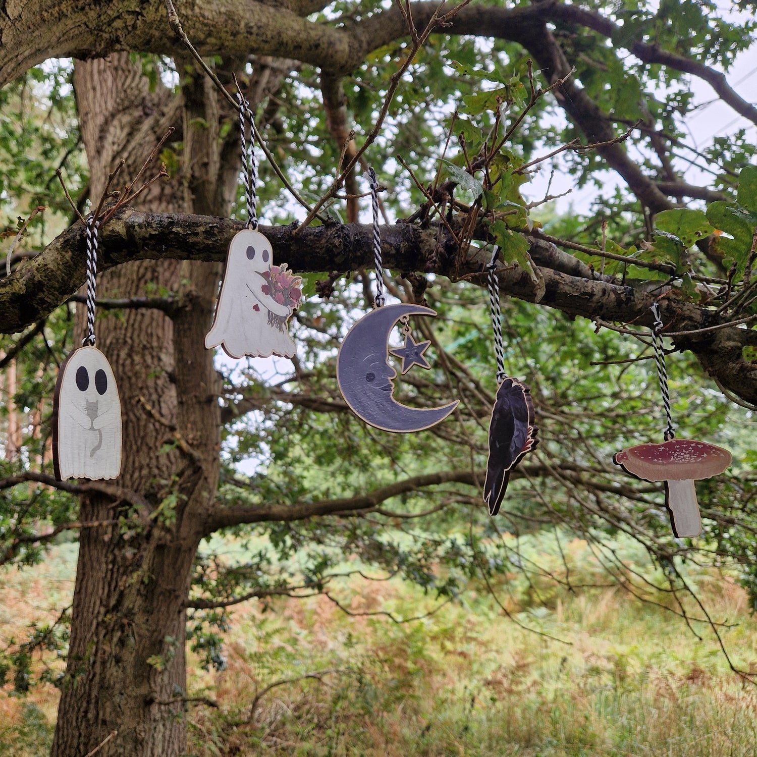 Five wooden charm decorations hanging from a tree in the woods. From left to right - a ghost holding a cat, a ghost holding some flowers, a crescent moon with a star hanging from it, a black fluffy pigeon then a red fly agaric toadstool.