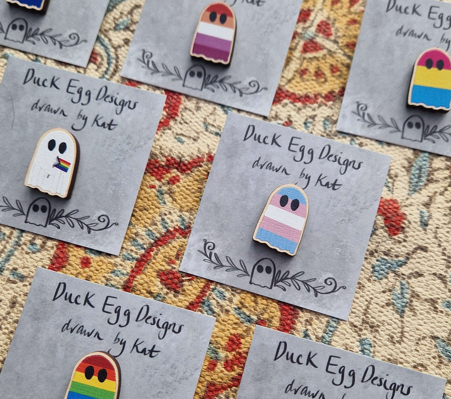 Pride flag ghost pin badges on grey backing cards sit on a floral patterned fabric.