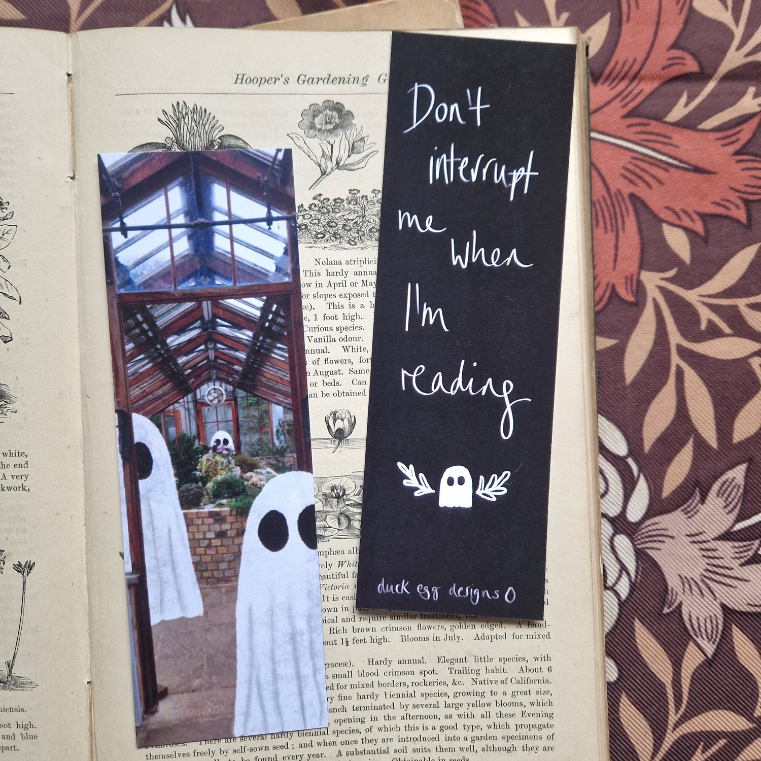 Two bookmarks on the open pages of an old book showing the front and back of the design. The left bookmark shows a greenhouse with ghosts while the write is black with the words 'Don't interrupt me when I'm reading'.