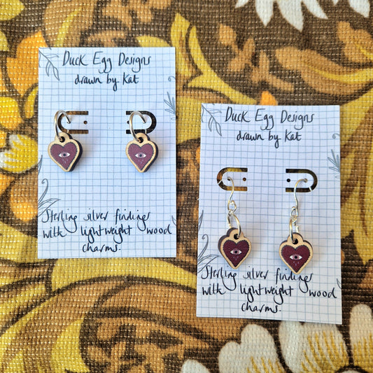 Two pairs of Mini Heart Dangle Earrings from Duck Egg Designs, with purple wooden charms and sterling silver accents, rest on display cards with handwritten notes atop floral fabric—an ideal Valentine’s Day gift.