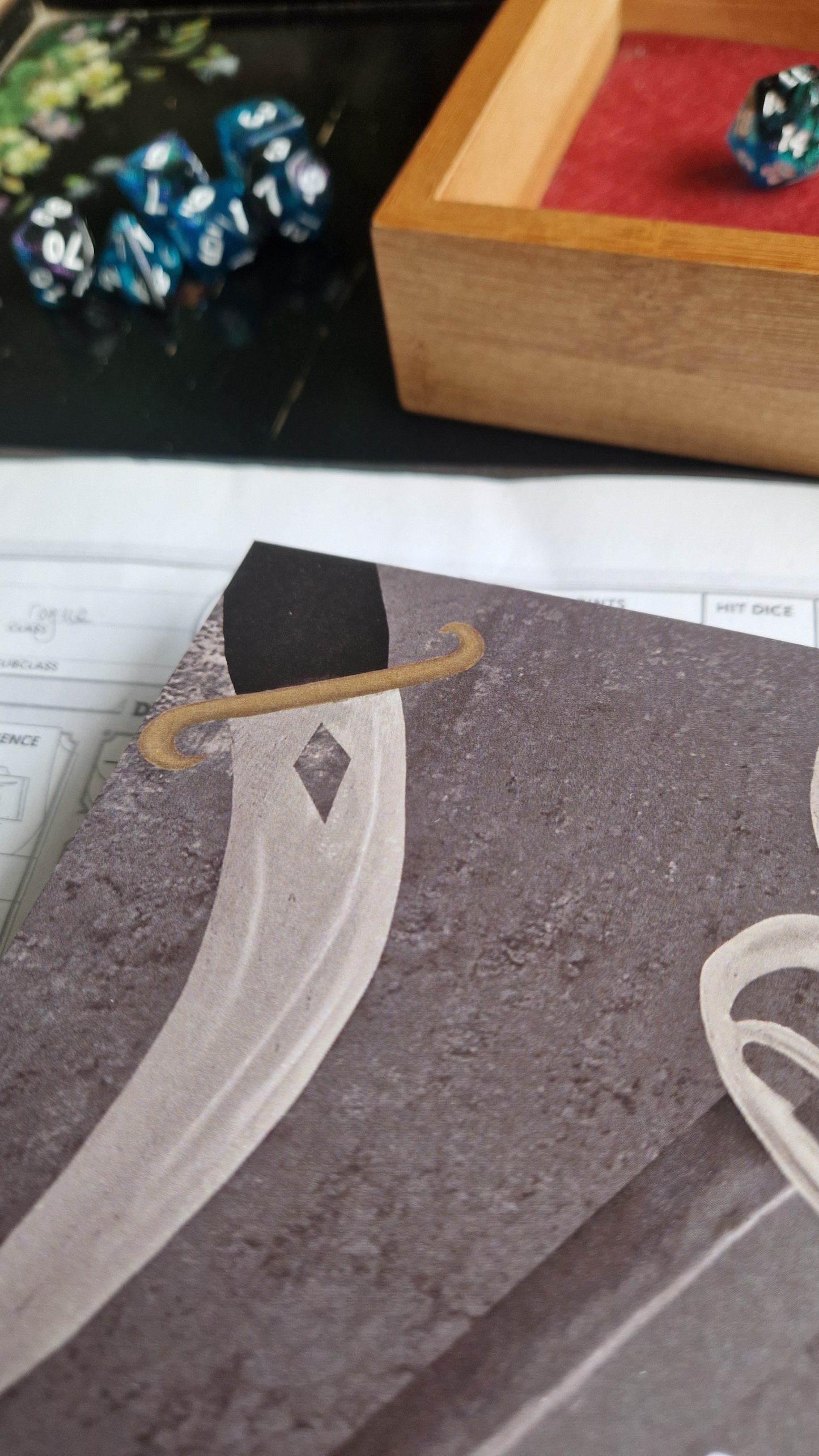 A close-up of a tabletop gaming setup featuring polyhedral dice, a red dice tray, campaign notes on recycled paper, a character sheet, and the Duck Egg Designs Cunning Actions Notebook with a stylized sword on the cover.