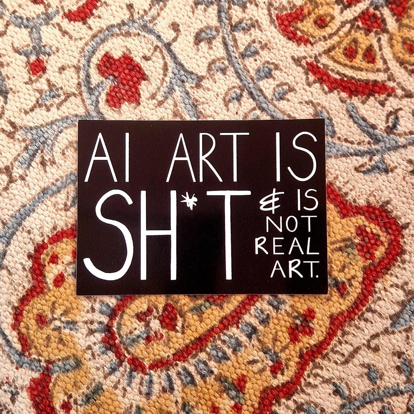 A black rectangular sticker featuring the words 'AI ART IS SHIT & IS NOT REAL ART. ' in white handwriting. The sticker sits on a patterned fabric background.