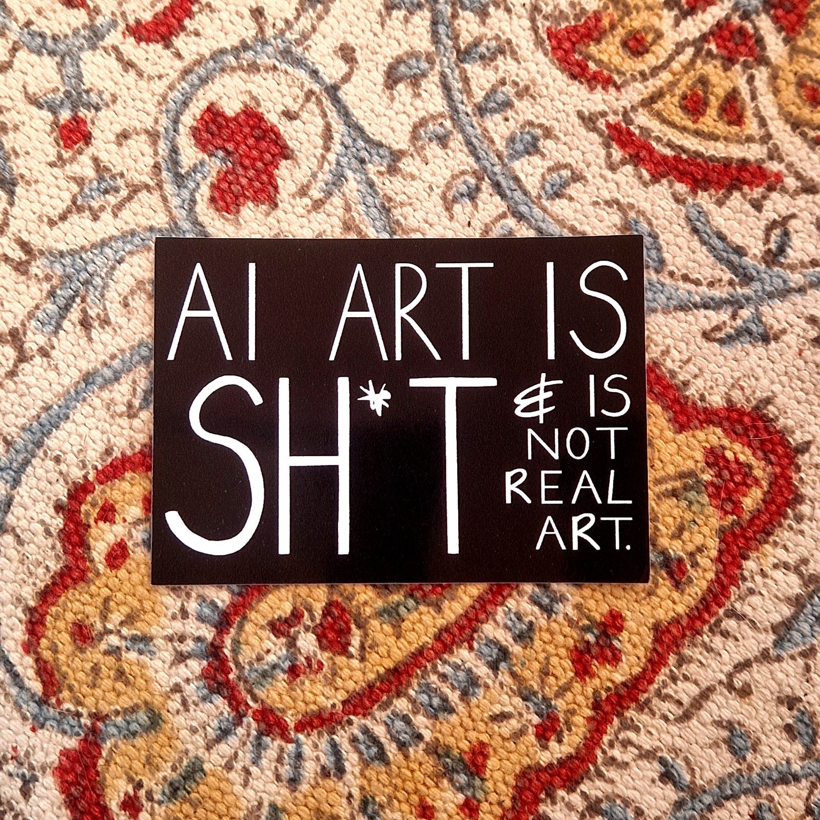 A black rectangular sticker featuring the words 'AI ART IS SHIT & IS NOT REAL ART. ' in white handwriting. The sticker sits on a patterned fabric background.