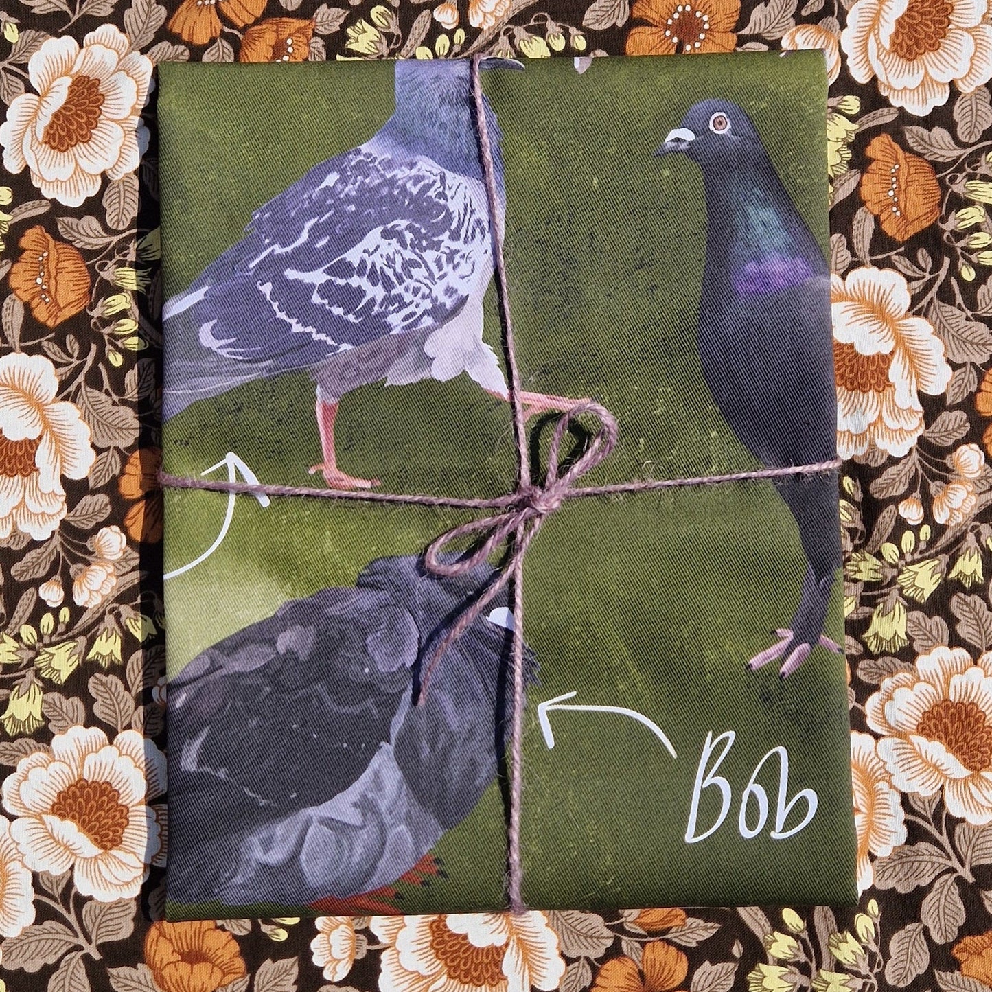  A green folded teatowel featuring pigeons sits on a retro floral patterned fabric. The teatowel is wrapped in a purple twine.