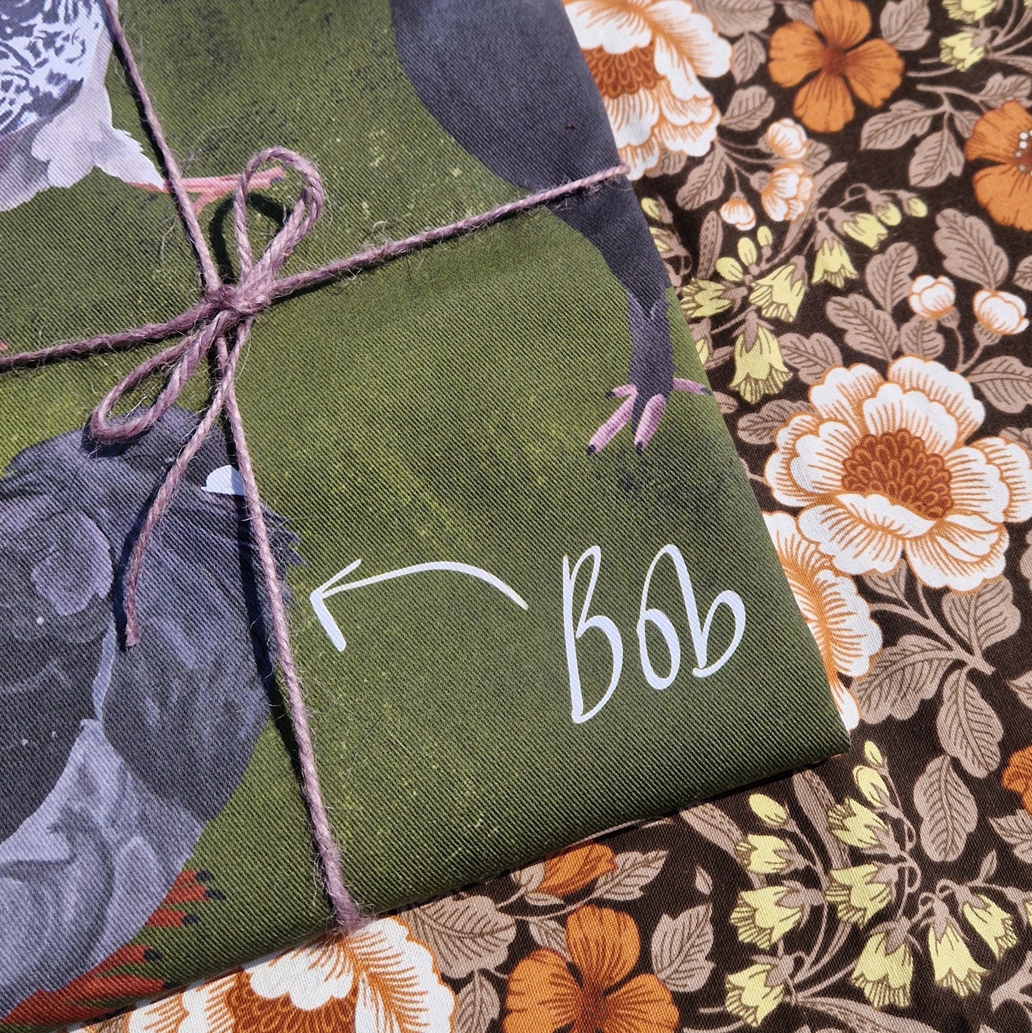A close up of the folded teatowel showing the detail in the illustrations. The teatowel is wrapped in a purple twine and sitting on a retro floral background.