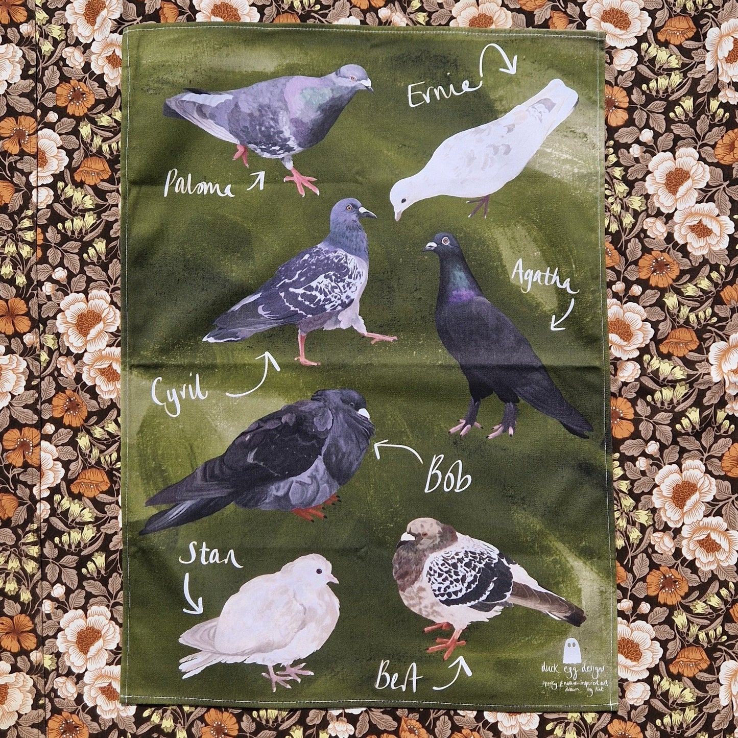 A green teatowel featuring seven different pigeons with their names in white handwriting next to them. Behind the teatowel is a retro floral patterned fabric.