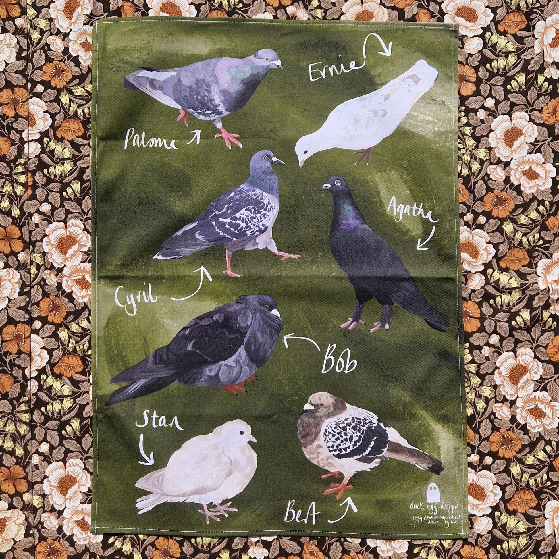 A green teatowel featuring seven different pigeons with their names in white handwriting next to them. Behind the teatowel is a retro floral patterned fabric.