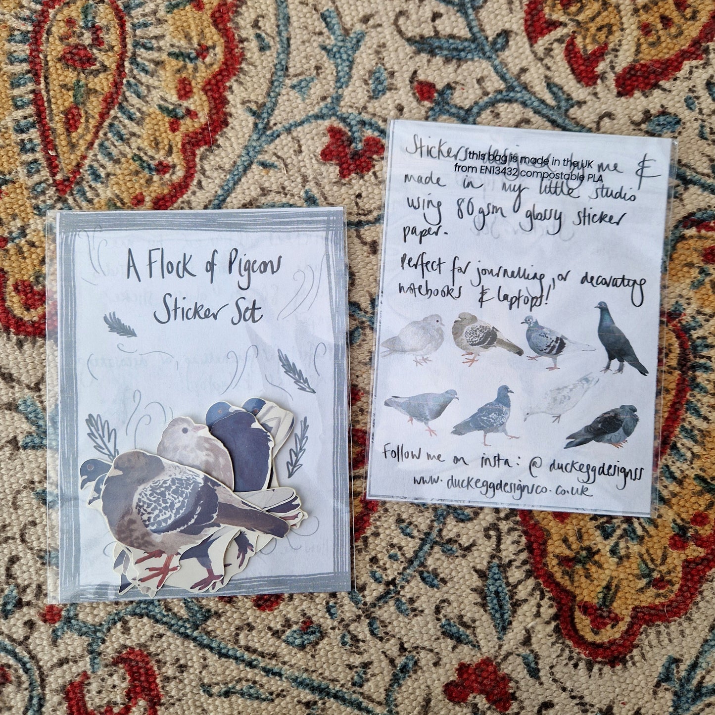 Two sticker sets next to each other - one with the top of the sticker set facing upward showing the stickers inside and one face down showing the sticker set information and the illustrations of all the stickers inside. They're both sat on a patterned fabric background.